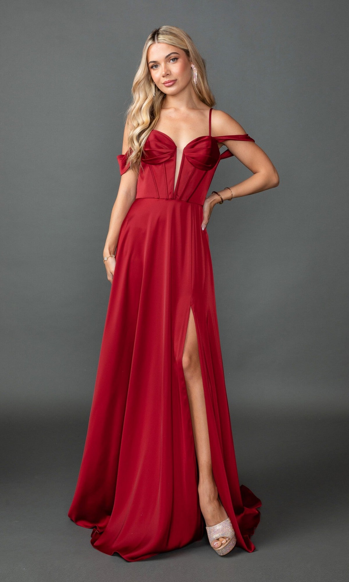 A woman poses against a plain gray background wearing the Long Prom Dress: Dancing Queen 4544—a red, floor-length A-line gown with spaghetti straps, off-the-shoulder sleeves, deep neckline, and thigh-high slit.